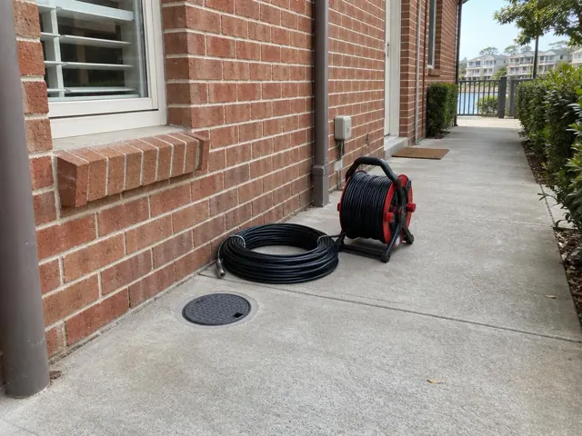 Flush round drain inspection cover beside a coiled camera reel on a brick townhouse walkway in Breakfast Point