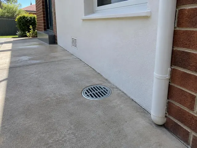 Concrete patio with a round metal gully grate beside a house wall and brick corner in Homebush